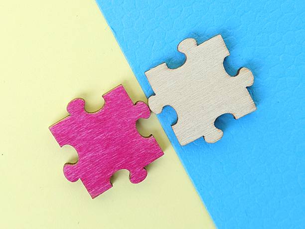 Two interlocking puzzle pieces, one pink and one natural wood, sit on a diagonal yellow and blue surface. The image evokes connection, teamwork, problem solving and creative collaboration