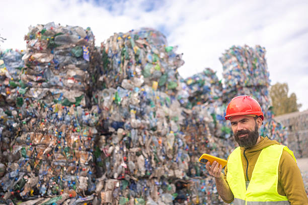 istock Bearded Man In Safety Gear Checking Mobile Phone Near Large Stacks Of Compressed Plastic Bottles At Recycling Plant 2245672302