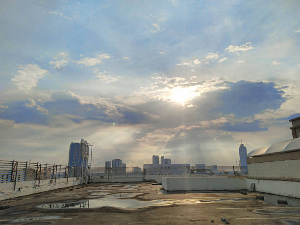 A dramatic cityscape view of Jakarta, Indonesia, taken from a rooftop after rain. The scene captures sunlight piercing through thick clouds, creating soft beams and reflections on puddles across the rooftop surface. The image conveys calmness, renewal, and beauty after the rain, suitable for themes of weather, inspiration, urban lifestyle, and architecture.
