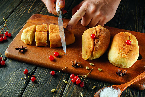istock Hands hold a knife as they slice warm bread rolls on a wooden cutting board. Bright red cherries and various spices are scattered around, adding color and flavor 2245709117