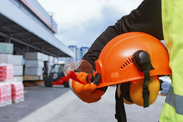 istock Orange construction helmet with earmuffs and safety goggles held by a worker in high-visibility gear at a warehouse site. Industrial safety, personal protective equipment in logistics. 2245727785
