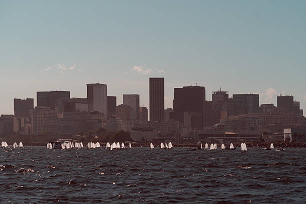 Telephoto view of Rio de Janeiro's Gloria district skyline with plenty of small white sport sailboats silhouettes gliding on dark ocean waves under warm light, blending urban and maritime atmosphere