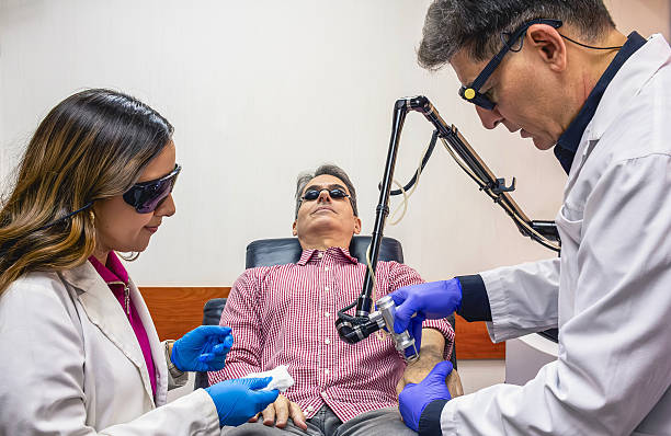 A male patient lies back while two doctors, wearing gloves and goggles, work on his forearm with laser device, demonstrating a specialized dermatology or cosmetic application beyond just the face.