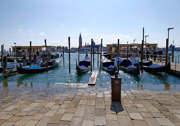 Traditional Venetian gondolas lined up on calm water near a wooden pier, with a scenic view of the lagoon and boats under a dramatic sky.