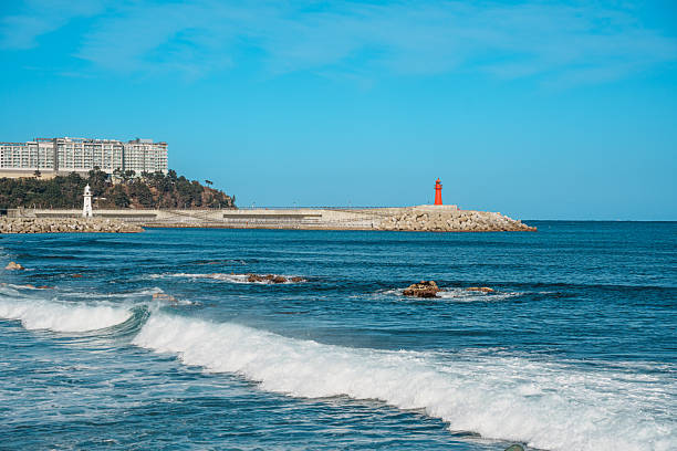 Waves roll toward the breakwater at Sokcho Pier, South Korea, with re lighthouses under clear blue sky.