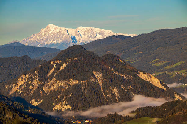 Austrian Alps landscape with forested hills and distant snow covered peaks under blue sky