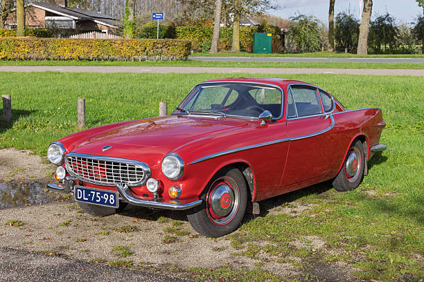 Front-side view of a beautifully preserved classic red Volvo P1800 coupe parked on the grass and gravel, featuring Dutch license plates. Utrecht Netherlands, 2 November 2025.