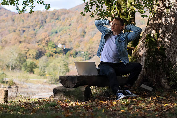 istock Man stretches arms behind head relaxing with laptop in fall scenery 2245833609