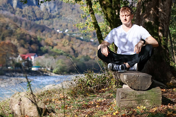 istock Man with eyes closed sitting on log by tree facing river valley 2245833839