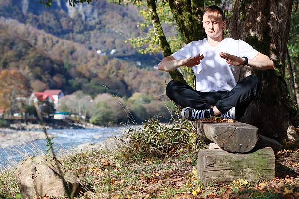 istock Man cross-legged on log near tree holding arms out with palms open facing river and mountain landscape 2245833862