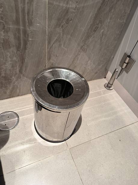 istock A high-angle shot of a cylindrical, stainless steel open-top trash bin standing on a light gray tiled floor in a modern restroom. The bin has a black liner visible inside and is positioned against a wall covered in dark gray marble-patterned tiles. 2245836097