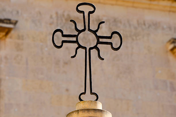 A close-up of a beautifully crafted wrought iron cross mounted on a stone pillar. The elegant curves and handcrafted design stand out against the warm stone background, symbolizing faith, heritage, and timeless artistry in metalwork.