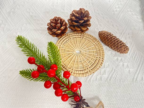 Natural holiday decorations featuring pinecones, red berries, and a woven coaster arranged on textured white fabric background for festive ambiance