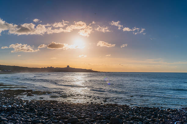 Summer sunset in Shimen District, New Taipei City, Taiwan, with golden sunlight reflecting on calm sea, rocky shore, and distant hill silhouette.