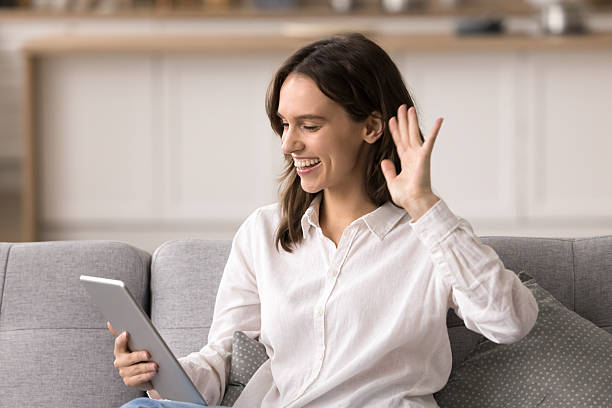 istock Friendly smiling millennial woman waving hand using tablet on sofa 2245867656