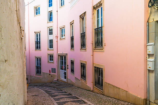 A quiet alleyway lined with cobblestones curves past a pastel pink building featuring wrought iron window railings and multiple balconies.