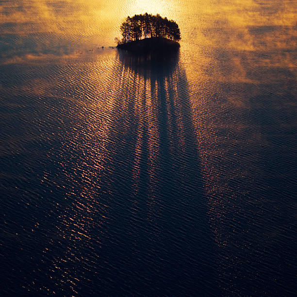 A small island with of trees casting shadows from the morning sunlight on the surface of misty lake.