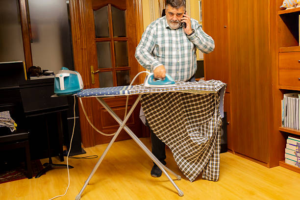 Mature man performs domestic chores, ironing clothes and making a phone call at home