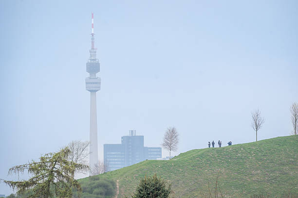 Urban Dortmund landscape with communication tower and silhouettes of people on grassy hill under soft blue sky.