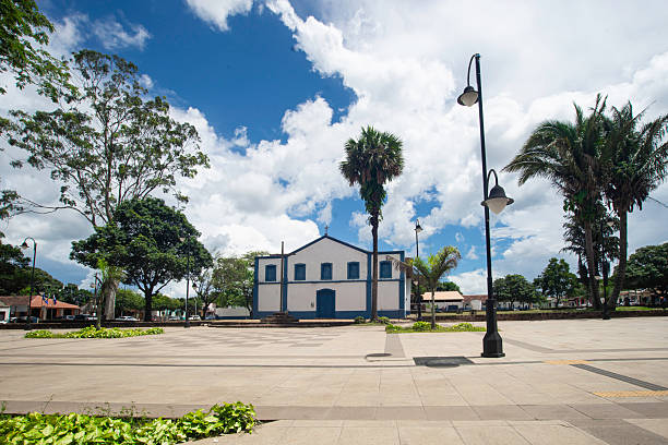 Panoramic view, church with a white facade and blue details, leafy trees to the side, a large cross on the sidewalk in front, a tall and slender palm tree, foreground, paved square with two garden areas, lamppost, background, low buildings, houses and shops, parked cars, city of Chapada dos Guimarães, blue sky with clouds