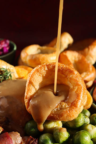 Close-up image Sunday roast dinner with all the trimmings, gravy being poured on Yorkshire pudding , roast chicken with crispy, golden skin, surrounded by side dishes of Brussel sprouts, roast potatoes, pigs in blankets, focus on foreground