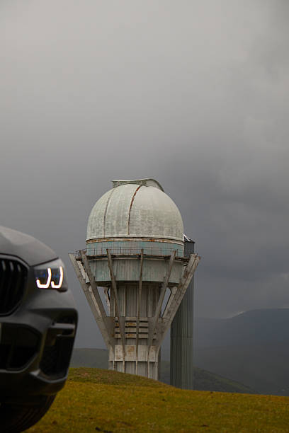 Naryn, Kyrgyzstan - May 13, 2024: Close-up vertical shot of the tall, weathered observatory dome structure, with the front of a BMW X5 G05 blurred in the foreground under a dark, stormy sky