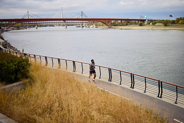 A lone man jogs along a riverside path with a red bridge in the distance.