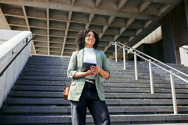 A confident young African American businesswoman is standing halfway up a large stone staircase, holding a digital tablet and smiling, looking forward with ambition and success.