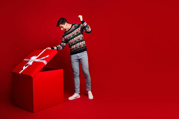 istock Playful man opens large red gift box in a festive Christmas scene wearing a patterned sweater jeans and white sneakers 2245966058