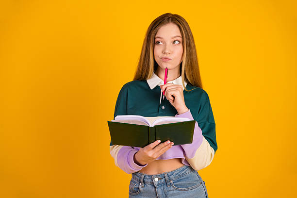 istock Young student holding a book, thinking while standing in front of a yellow background in casual wear 2245966818