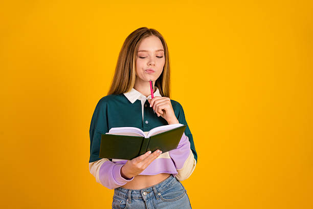 istock Lovely female student thoughtful while reading a notebook against a yellow background, wearing trendy casual attire 2245966828