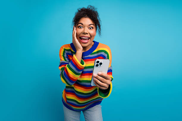 istock Young woman with colorful striped sweater surprised while using a smartphone against a blue background for lifestyle and fashion marketing 2245967112
