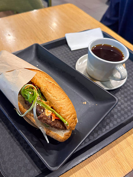 Savoring a delicious, fresh grilled meat Banh Mi sandwich with vibrant herbs and a hot cup of black coffee, served casually on a tray. Ideal quick meal or snack.