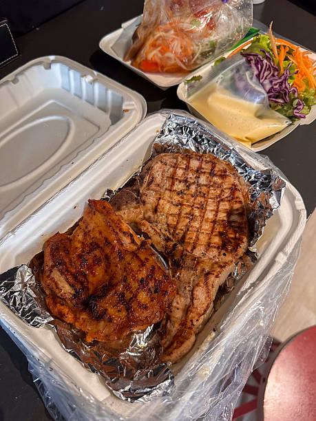 Delicious grilled pork chop and chicken, expertly prepared and packaged in a takeout container with foil, accompanied by fresh Thai papaya salad and garden greens. Perfect for a convenient and