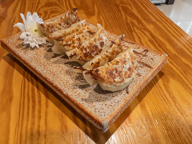 Delicious pan-fried gyoza dumplings beautifully arranged on a speckled ceramic plate with a white flower garnish, set against a warm wooden table. A perfect culinary delight.