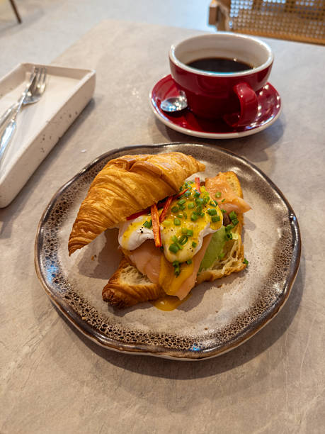 Delicious gourmet croissant sandwich featuring smoked salmon, creamy avocado, perfectly poached eggs with rich hollandaise, topped with chives, served with a hot coffee on a cafe table, ideal for