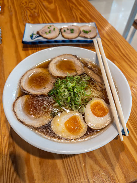 A steaming bowl of authentic Japanese ramen, featuring rich pork broth, tender chashu slices, perfectly marinated ajitama eggs, springy noodles, and fresh scallions, served on a rustic wooden table.