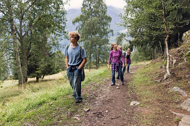 Group of hikers walk along a dirt path running on the side of lush forest. The hikers carry backpacks as walk up the hill. Shot with Canon R5 on an overcast summer day.