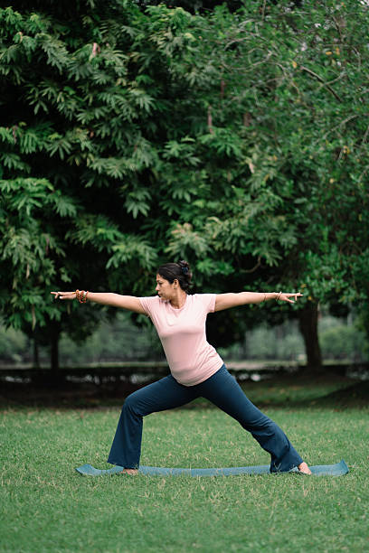 Full-length image of a woman performing the Warrior II (Virabhadrasana II) yoga pose on a mat in a lush green park. She is wearing a pink T-shirt and navy-blue pants, maintaining strength and balance while focusing on her posture. The scene represents mindfulness, body awareness, and a healthy lifestyle through outdoor yoga practice. Suitable for concepts related to wellness, fitness, meditation, and mental well-being.