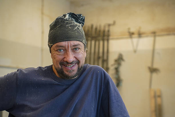 A middle-aged carpenter in his workshop, wearing a casual shirt and headband, smiling confidently in front of woodworking tools and machinery. The background captures the authentic atmosphere of craftsmanship and manual work. Perfect image for themes related to woodworking, craftsmanship, small business, and traditional trades.