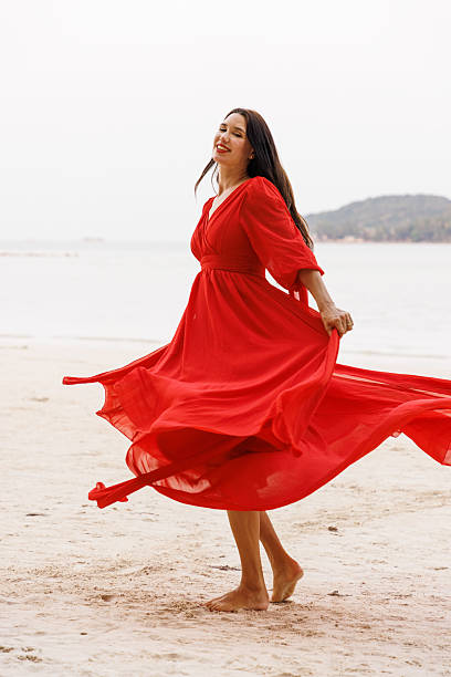 A smiling woman gracefully spinning in a vibrant red dress on a serene, sandy beach.