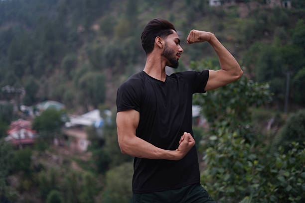 Man confidently flexing biceps in a black t-shirt with a lush green landscape backdrop. Ideal for fitness, lifestyle, and strength themes.