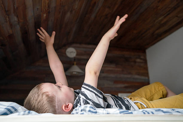 A young boy lies on a striped bedspread in a cozy attic, arms raised in playful stretch. The wooden ceiling and soft light create a warm, imaginative mood of exploration and joy.