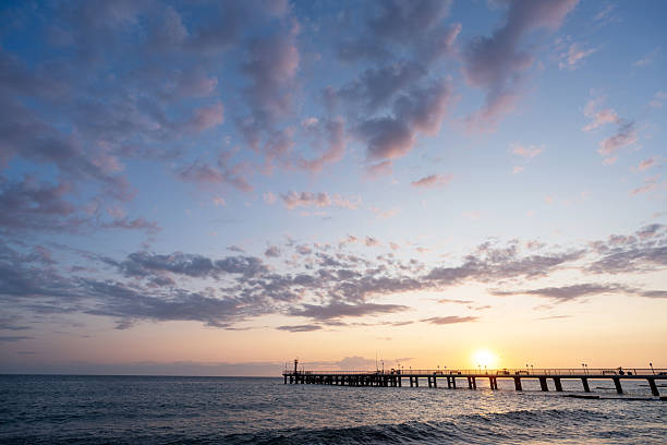 A serene sunset over the sea with a wooden pier reaching into the water. Warm light, soft clouds, and gentle waves create a peaceful, contemplative mood.