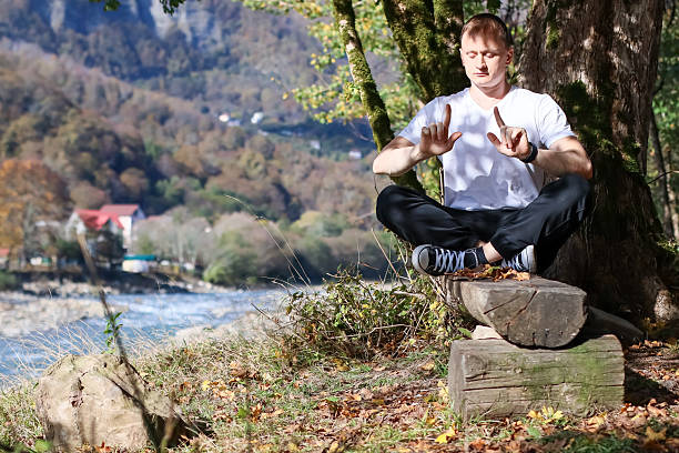 istock Man seated on log beside tree pointing fingers forward in meditation pose with mountain river background 2245984006