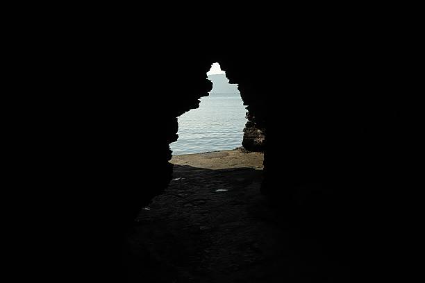 A dramatic low-key view from inside a dark, rugged cave, looking out through an irregular opening to a serene expanse of blue sea and distant horizon, highlighting the natural contrast between shadow and light.