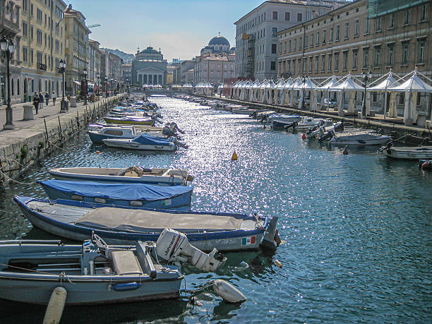 Trieste, Italy – 16 April 2011: Small boats float along the Grand Canal (Canal Grande) in central Trieste, with bright spring sunlight shimmering across the water. The surrounding architecture — dominated by neoclassical façades and orderly Austro-Hungarian urban planning — reflects the city’s long history under the Habsburg Empire, making Trieste visually distinct from typical Italian port towns. The canal, constructed in the 18th century to allow merchant vessels direct access to the city’s commercial heart, leads toward the Church of Sant’Antonio Taumaturgo, whose white neoclassical front and dome anchor this culturally layered Adriatic scene.