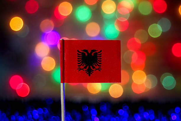 The national flag of Albania, featuring the iconic black double-headed eagle on a striking red field, is dramatically presented against an abstract, colorful, and luminous bokeh backdrop. This image powerfully conveys themes of Balkan identity, European pride, and historical resilience, ideal for celebrating Independence Day, major political events, or travel to the Adriatic Coast. The festive, multicolored lighting suggests joyful national festivals, New Year, or Christmas celebrations.
