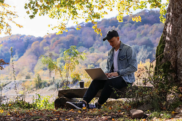 istock Young man types on laptop while sitting on bench in autumn forest with colorful hills 2245989992