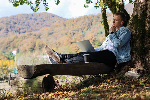 istock Young man reclining on log under tree with laptop in autumn mountain setting 2245990119